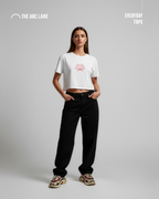 Woman wearing a white t-shirt with a pink lotus design and black pants on a gray background, branded 'The Arc Lane'.